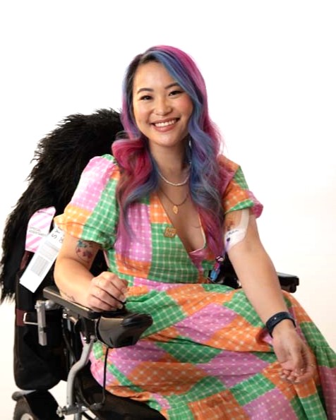Akii is smiling at the camera wearing a bright coloured dress. They have brightly coloured hair and they are sitting in a power wheelchair.