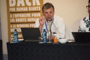 Dimitris Christopoulos sitting at table speaking to crowd in front of a laptop