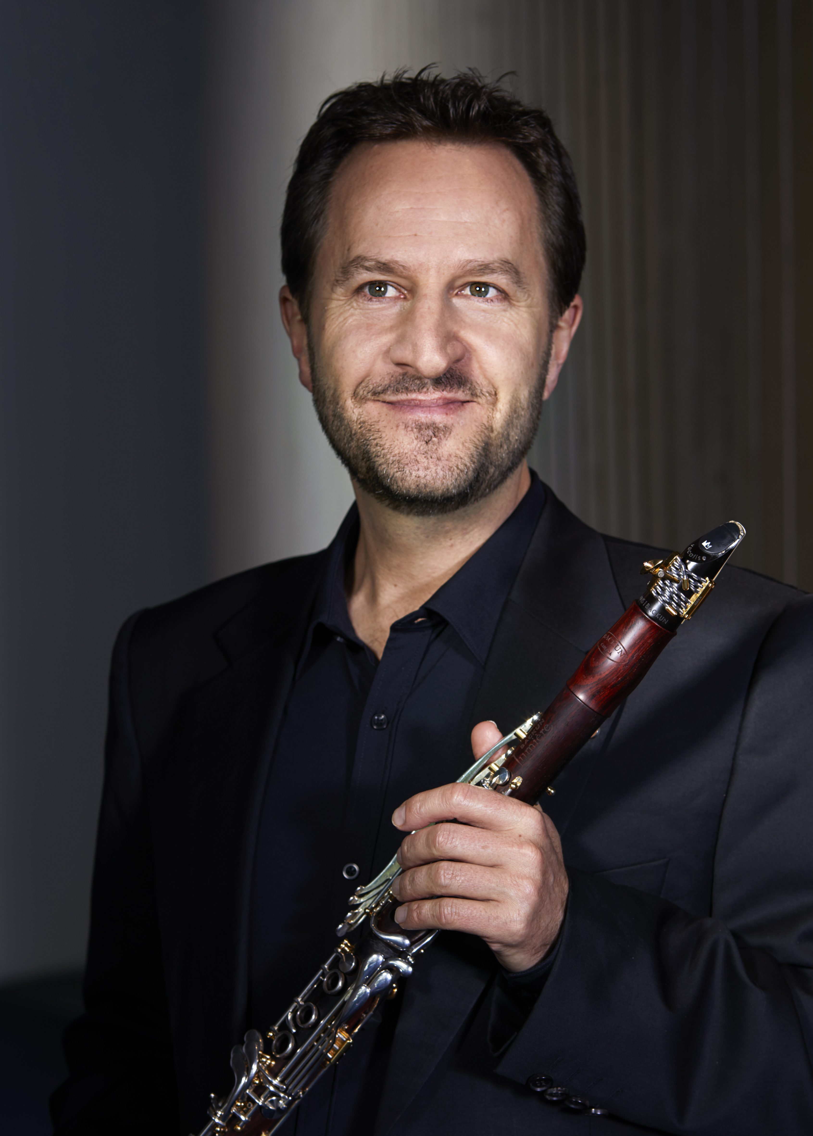 Man with short brwn hair and a short beard wearing a black suit and holding a clarinet