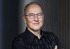 Ian Munro: A middle-aged man with short hair and glasses, wearing a black collared shirt smiles with his lips closed.