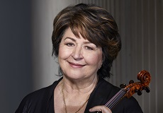 A woman with short brown hair wearing a black blouse and a necklace. She smiles with her lips closed and holds a viola.