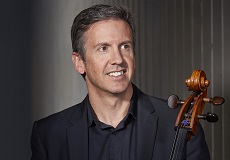 A man with short dark blonde hair, looks to the right of camera and smiles. He is weating a black collared shirt and a black jacket. He is holding a cello.