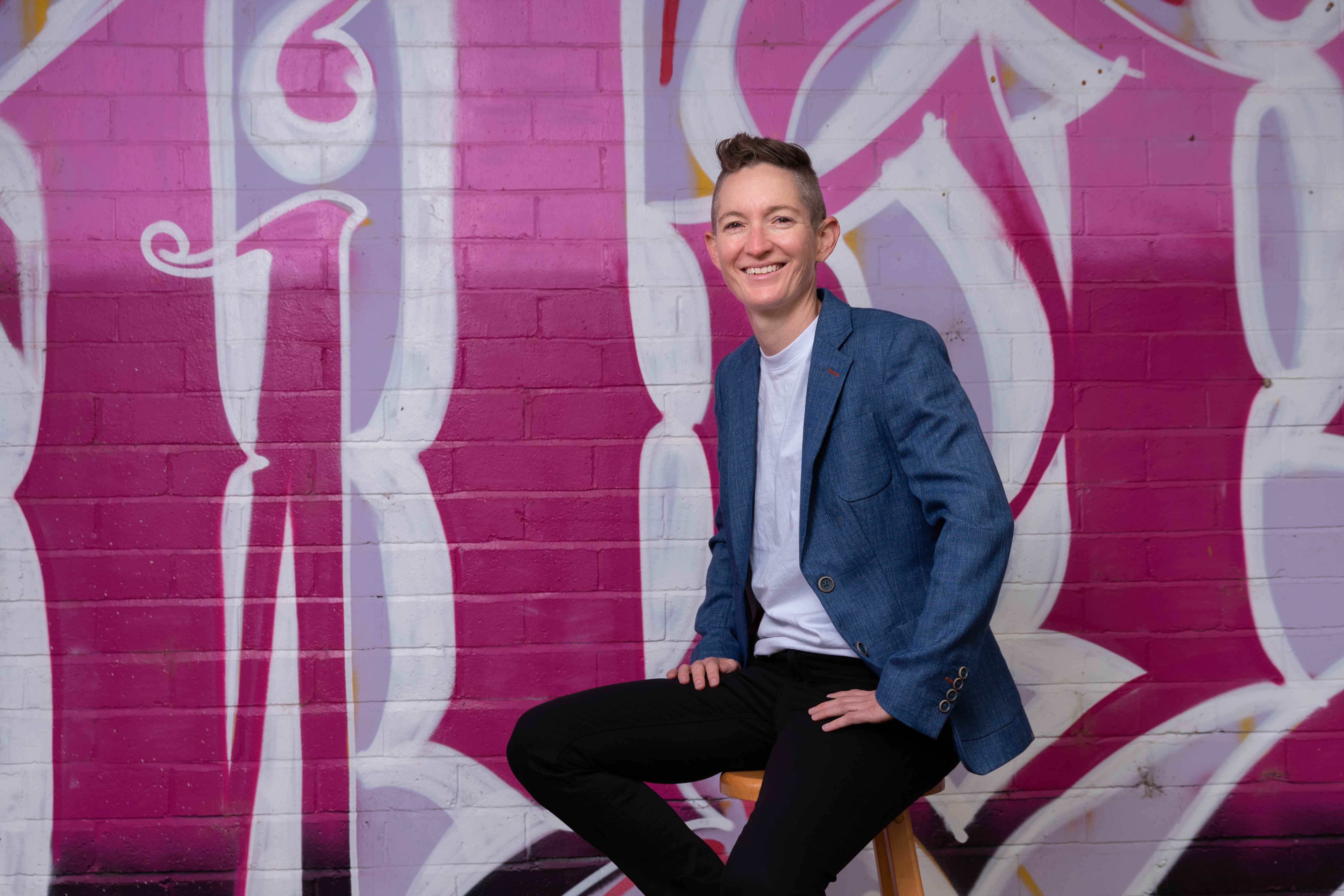 Bree is sitting on a stool in front of a wall that has been painted in pink graffiti. They are wearing a grey blazer and have short hair. They are smiling at the camera. 