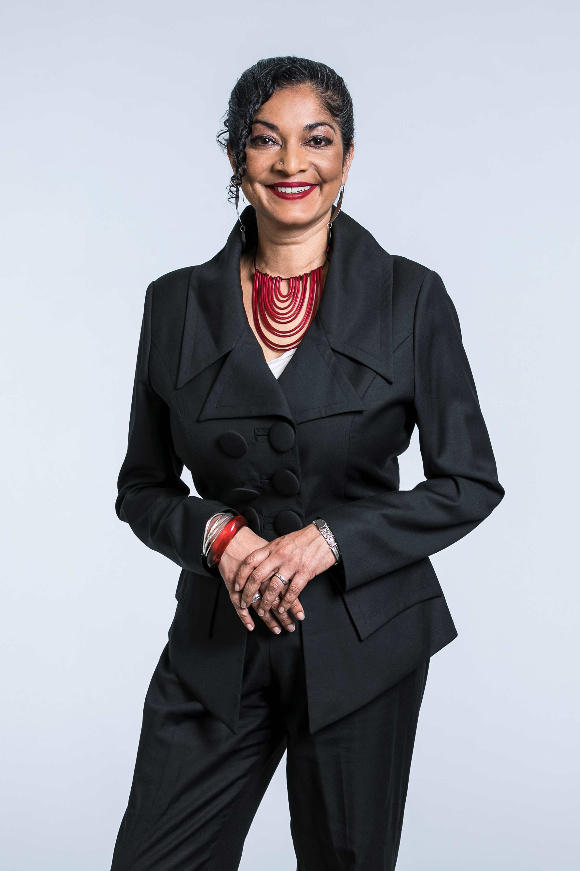 Padma is standing in front of the camera, smiling. She is wearing a black suit with a red statement necklace. Her black hair is tied back.