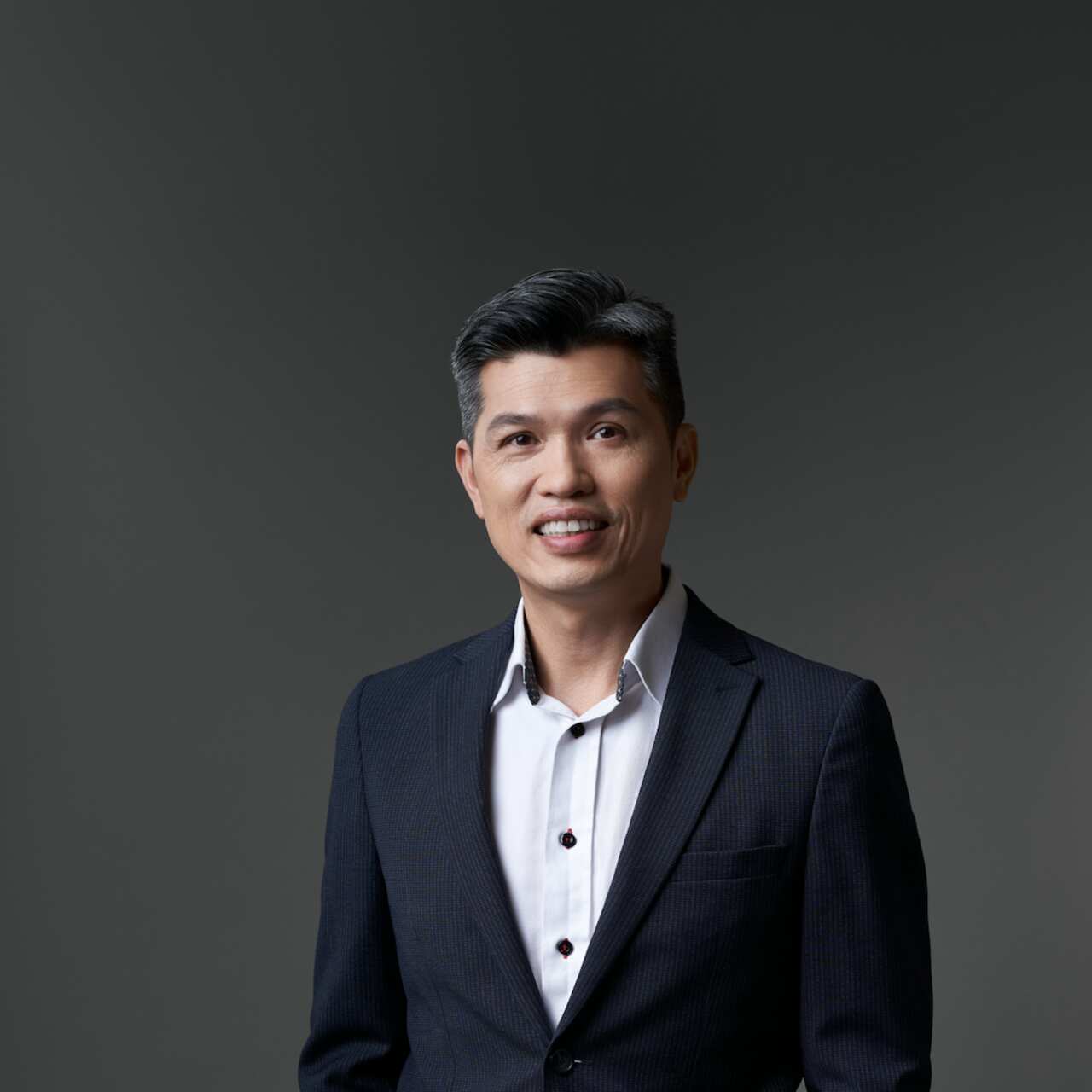 Tommy Lai smiles to the camera, he is wearing a black suit. 