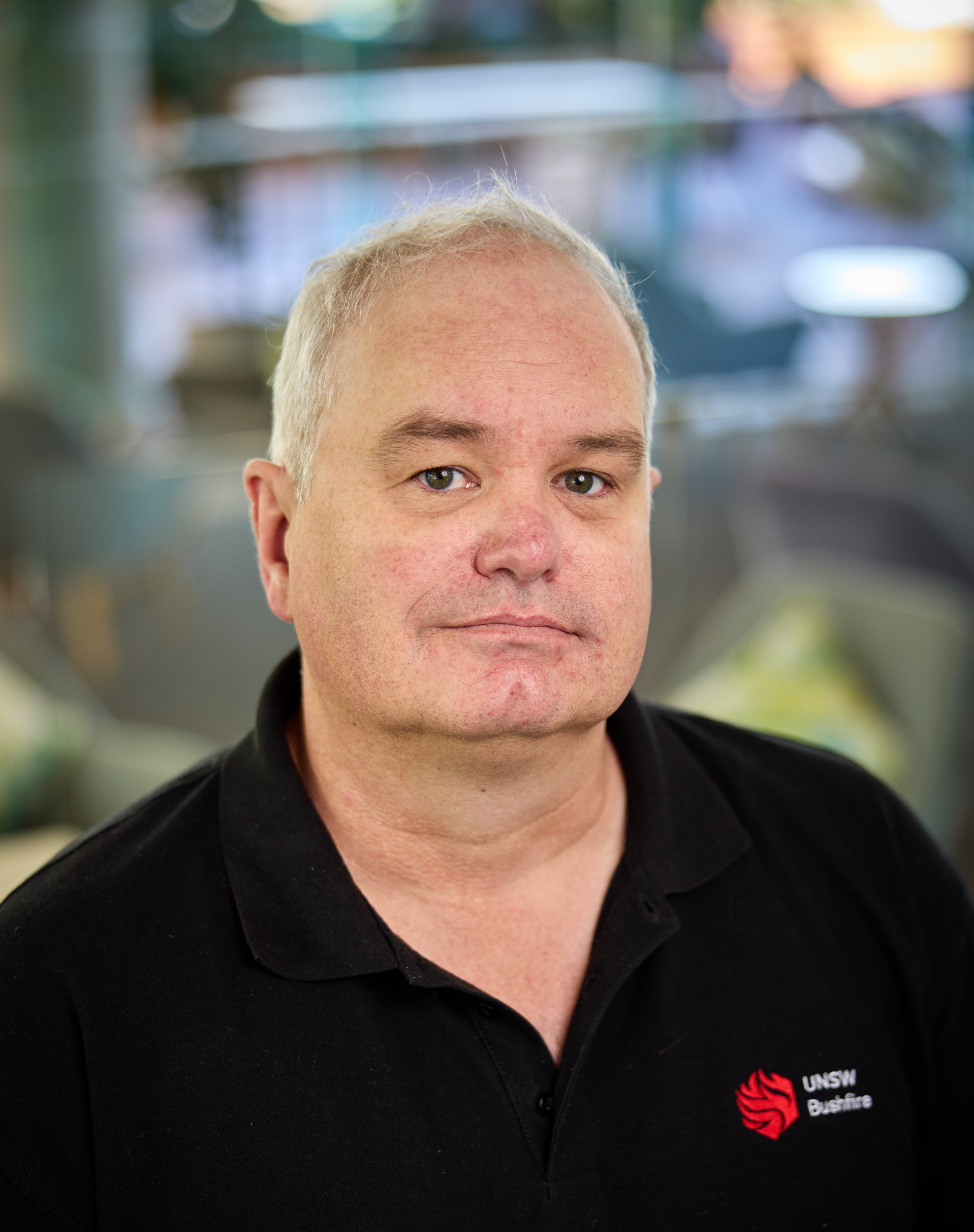 A man with brown eyes and white hair wearing a black polo with the UNSW Bushfire Research logo on the breast smiles slightly as he looks into the camera.
