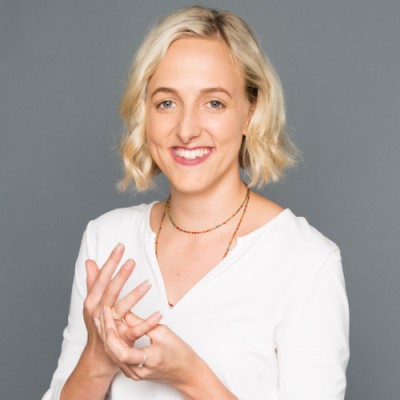 A woman with a wavy blonde bob haircut and brown eyes smiles towards the camera. She's wearing a white V-neck top with long sleeves, two gold necklaces (one short, one long), and appears to be fiddling with a ring on her left hand in the camera frame. 