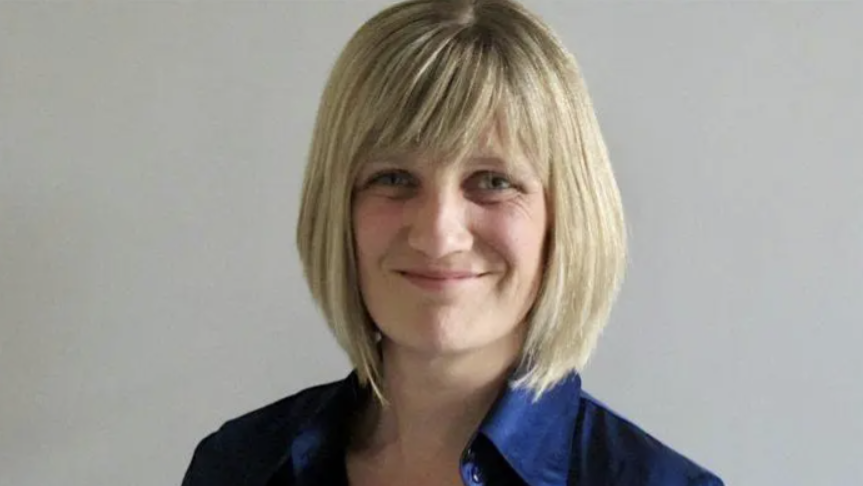 A smiling woman with a chin-length blonde bob wears a dark shirt with a bright blue collar against a plain light-gray wall.