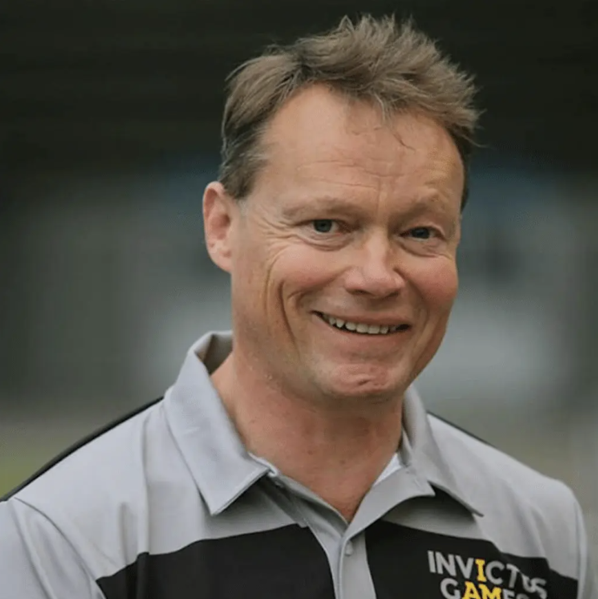 A smiling man wears a gray-and-black polo shirt bearing the Invictus Games logo against a softly blurred dark background.