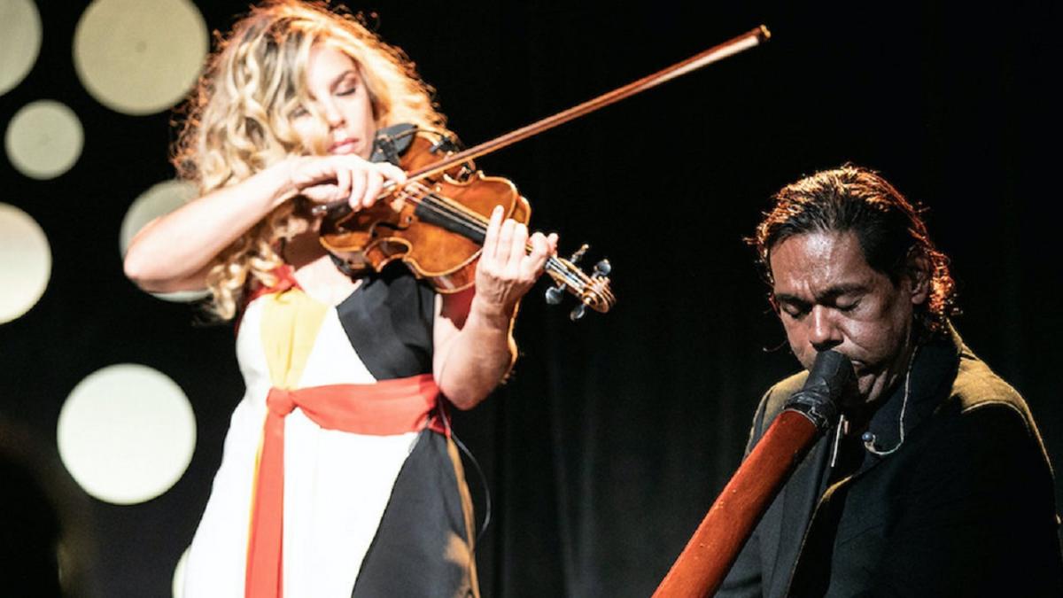 Woman with blonde hair playing violin next to a man sitting, playing the didgeridoo