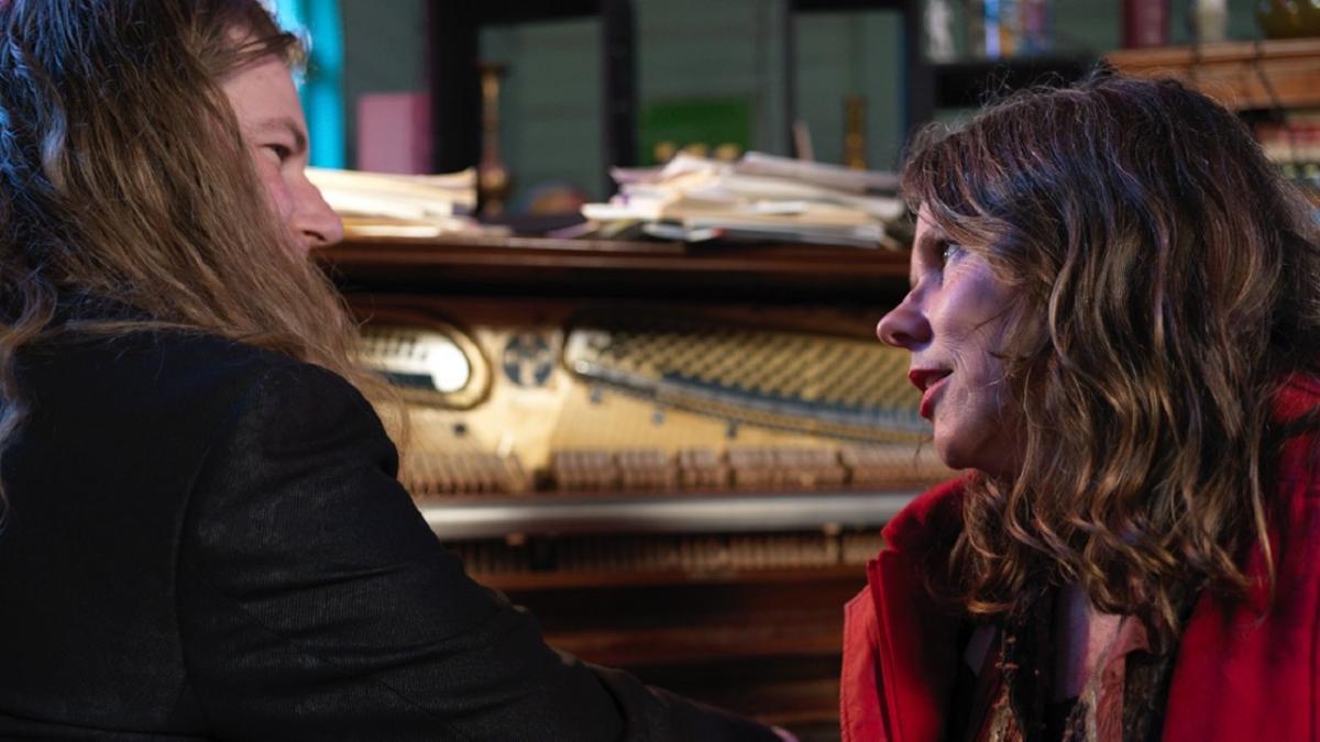 A man and a woman looking at each other, a piano in the background.