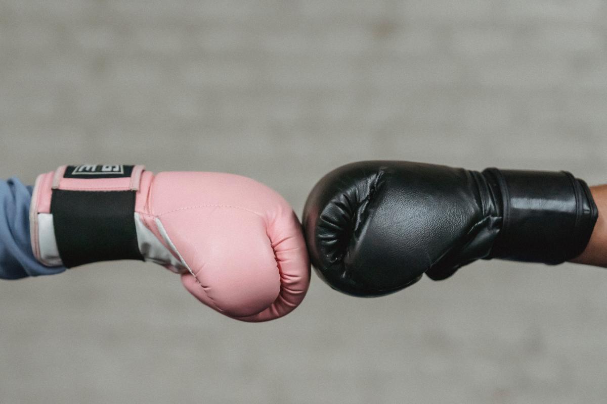 One pink and one black boxing glove meeting in the middle in front of a grey brick wall