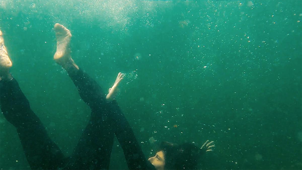 Photo of a man falling underwater, wearing a black wetsuit, bubbles all around him. 