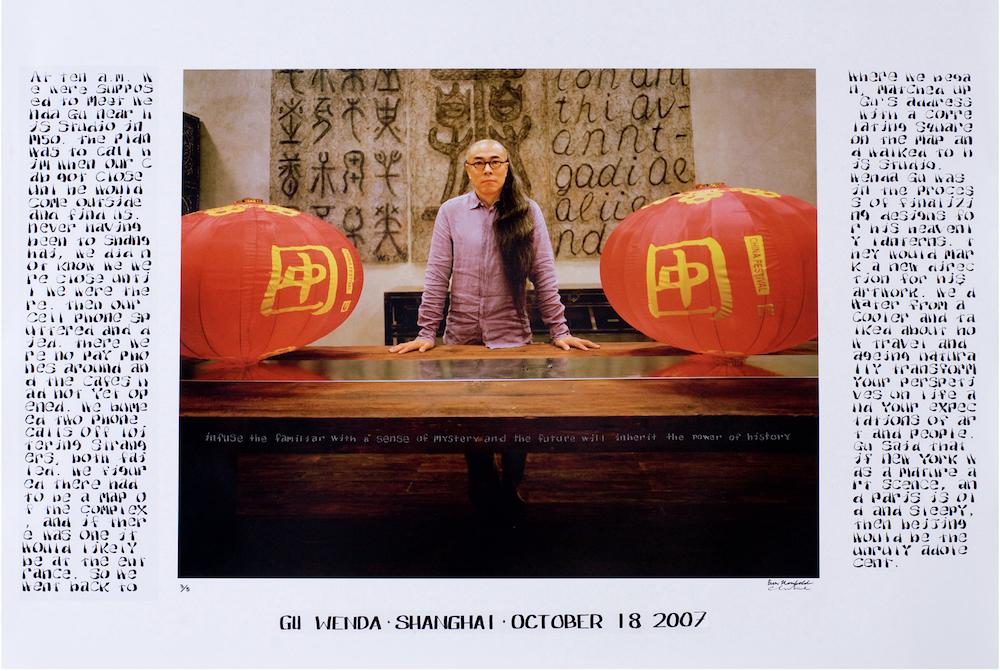 Image caption: Photographic portrait of artists Gu Wenda, standing at a table between two red lanterns. On either side of the photograph is a poem.