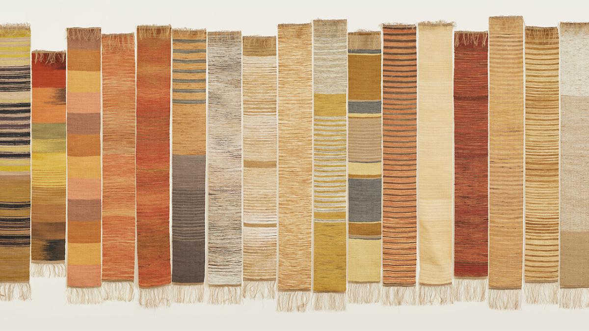 A row of 17 long, weavings hanging on a cream wall. The weavings have various horizontal striations in earthy colours.