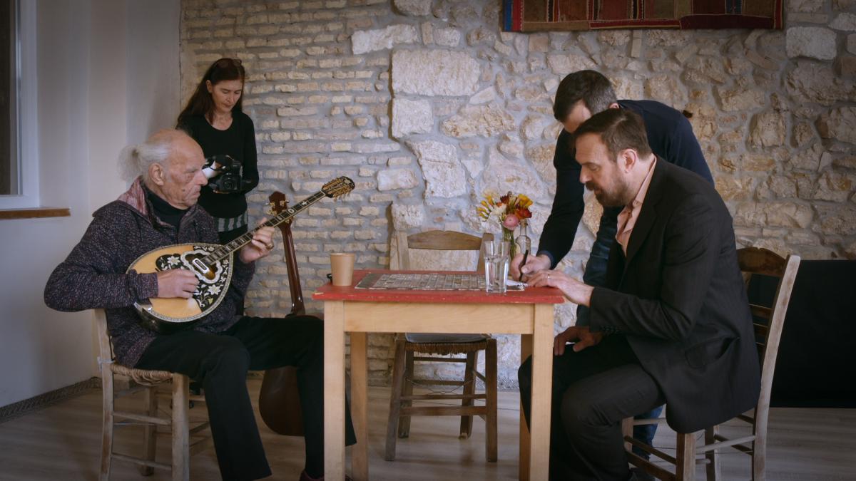 Four people around a small table in a stone walled room. One man is holding a stringed instrument as the man opposite him is writing. A woman is filming the men in the background. 