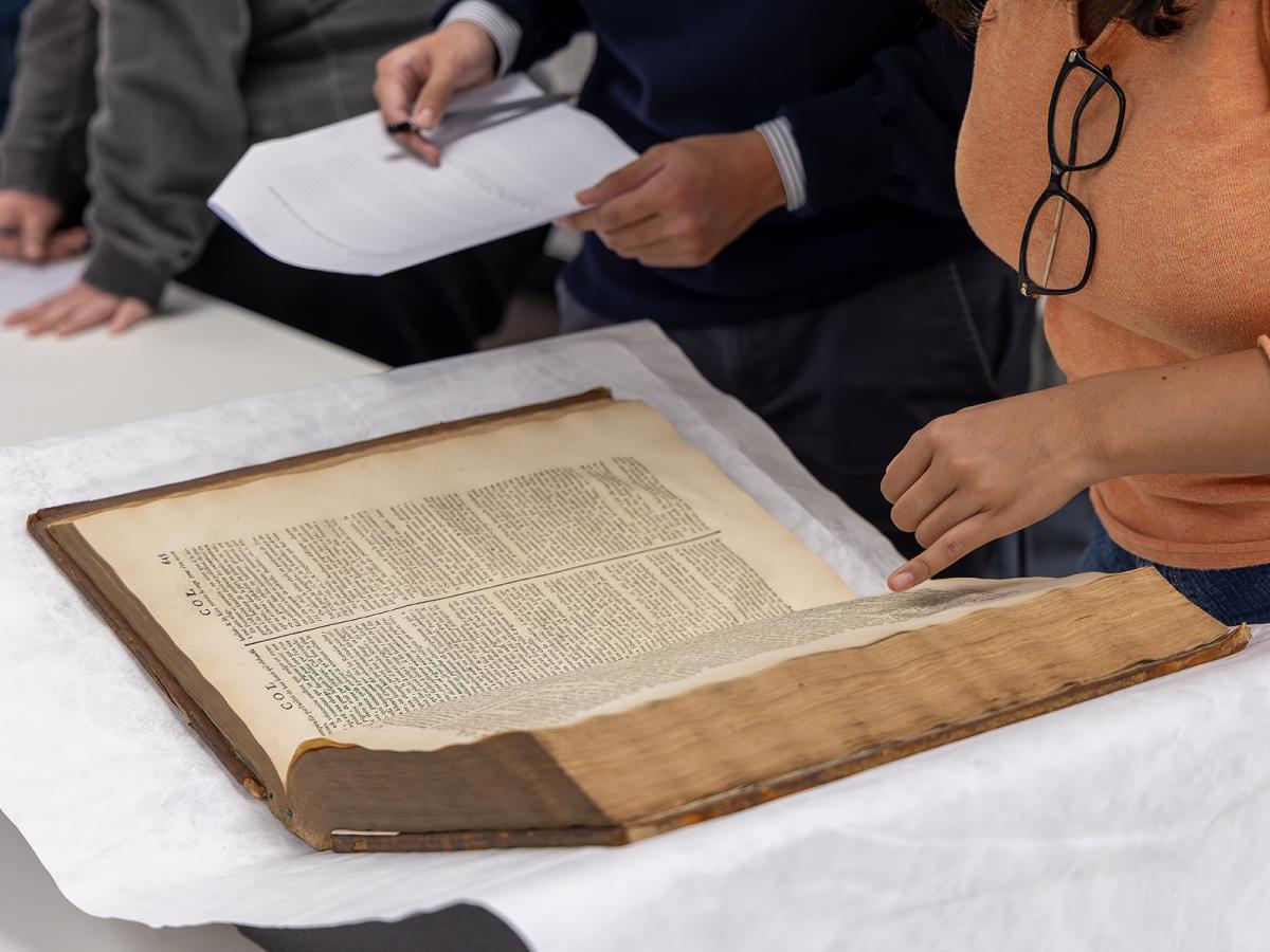 person in orange jumper standing in front of large antique looking book with pointed finger.