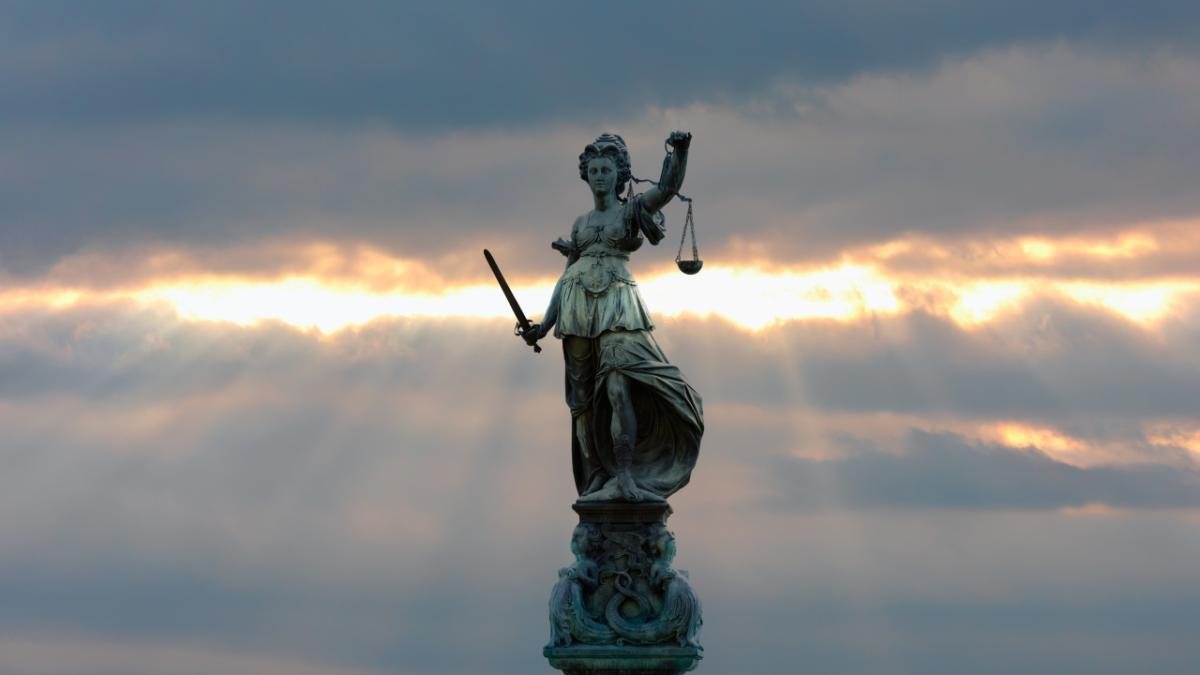 A centered, full-length bronze statue of Lady Justice stands on a dark, decorated pillar, wearing draped classical Roman robes.